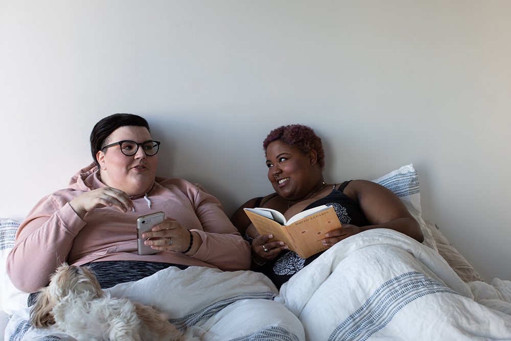 Two plus-sized femmes in bed together having friendly conversation. One is dark-skinned with short-cropped hair and is holding a book while the other is light-skinned with short-cropped hair and is holding their phone.