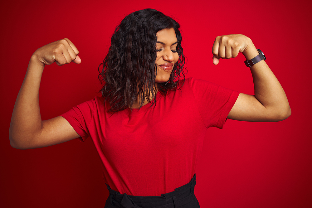 Beautiful trans woman wearing t-shirt over isolated red background showing arms muscles smiling proud.